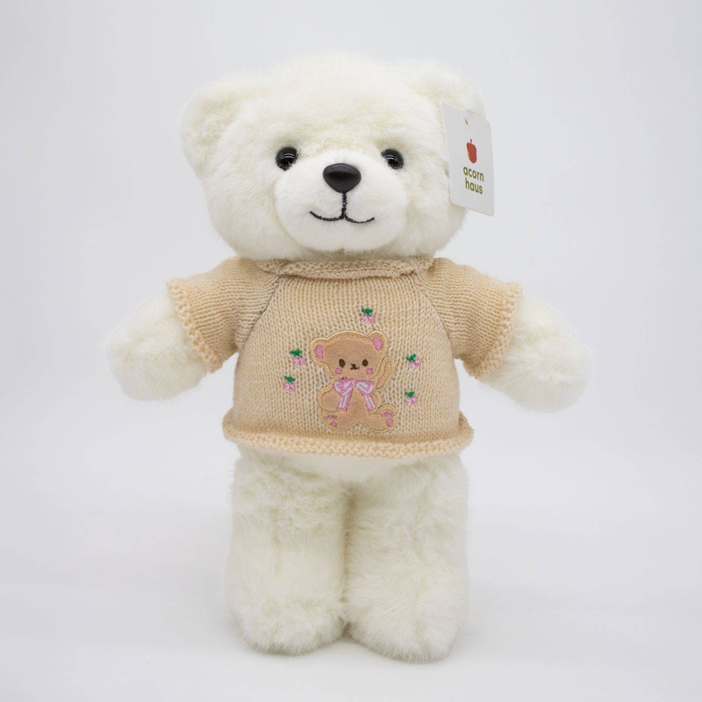 White Carrington teddy bear standing upright wearing a beige sweater with a brown bear wearing pink bow design on a white background