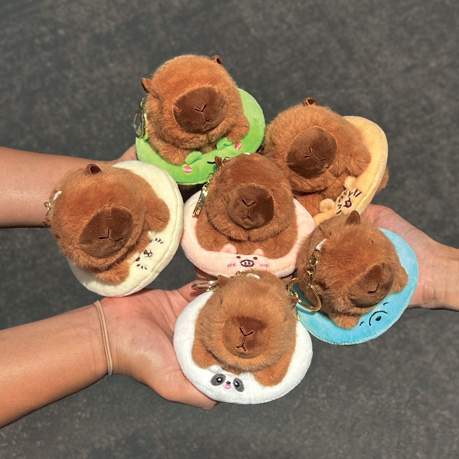 Three small plush capybara keychains or charms being held in three hands. Soft capybara keychain wearing a cute bear shaped backpack with a gold keyring keychain.