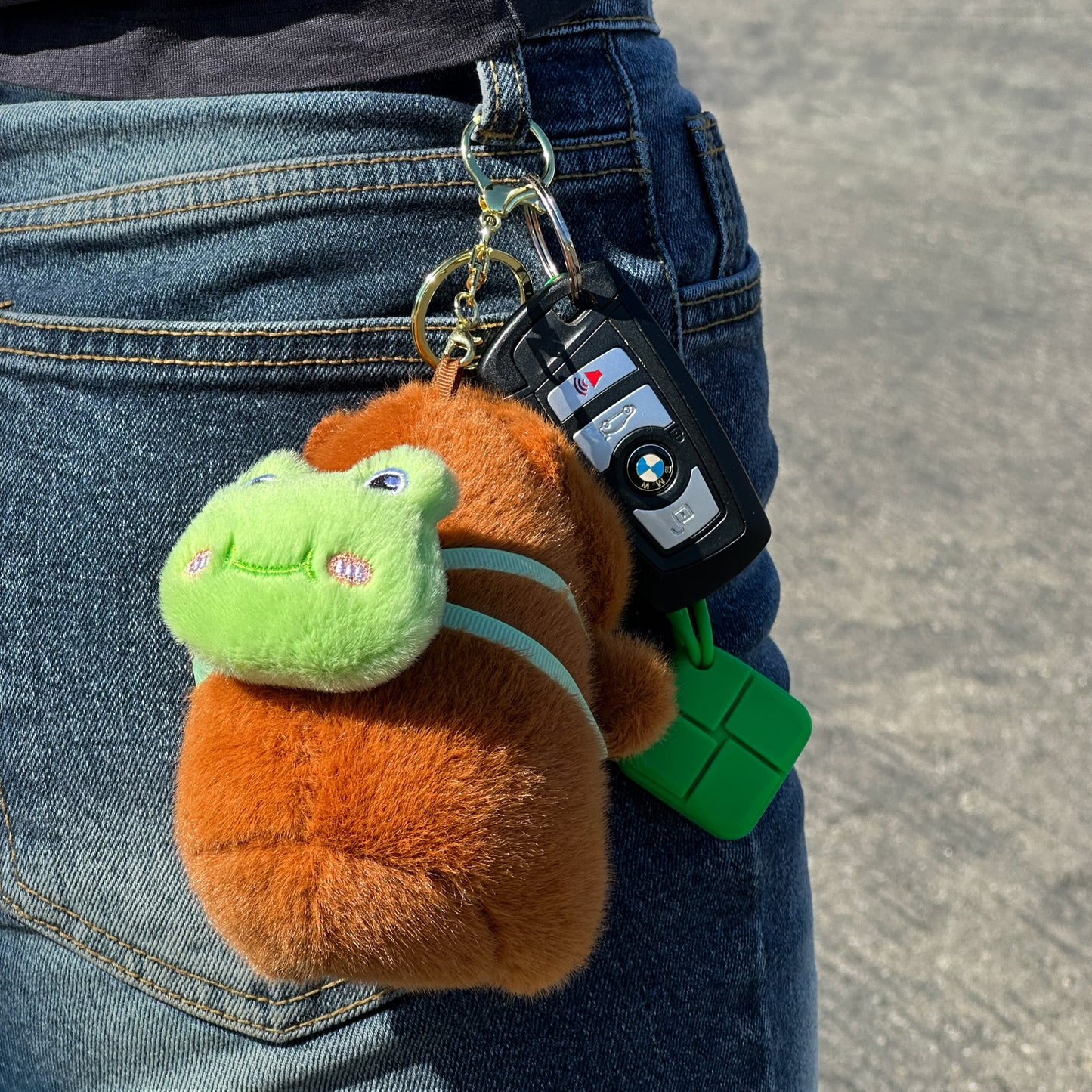 Soft capybara keychain wearing green frog backpack with a gold keychain hanging from jeans
