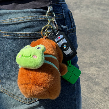 Soft capybara keychain wearing green frog backpack with a gold keychain hanging from jeans