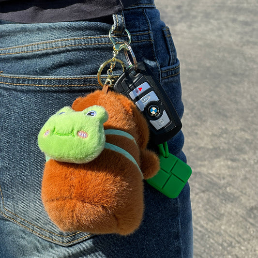 Soft capybara keychain wearing green frog backpack with a gold keychain hanging from jeans