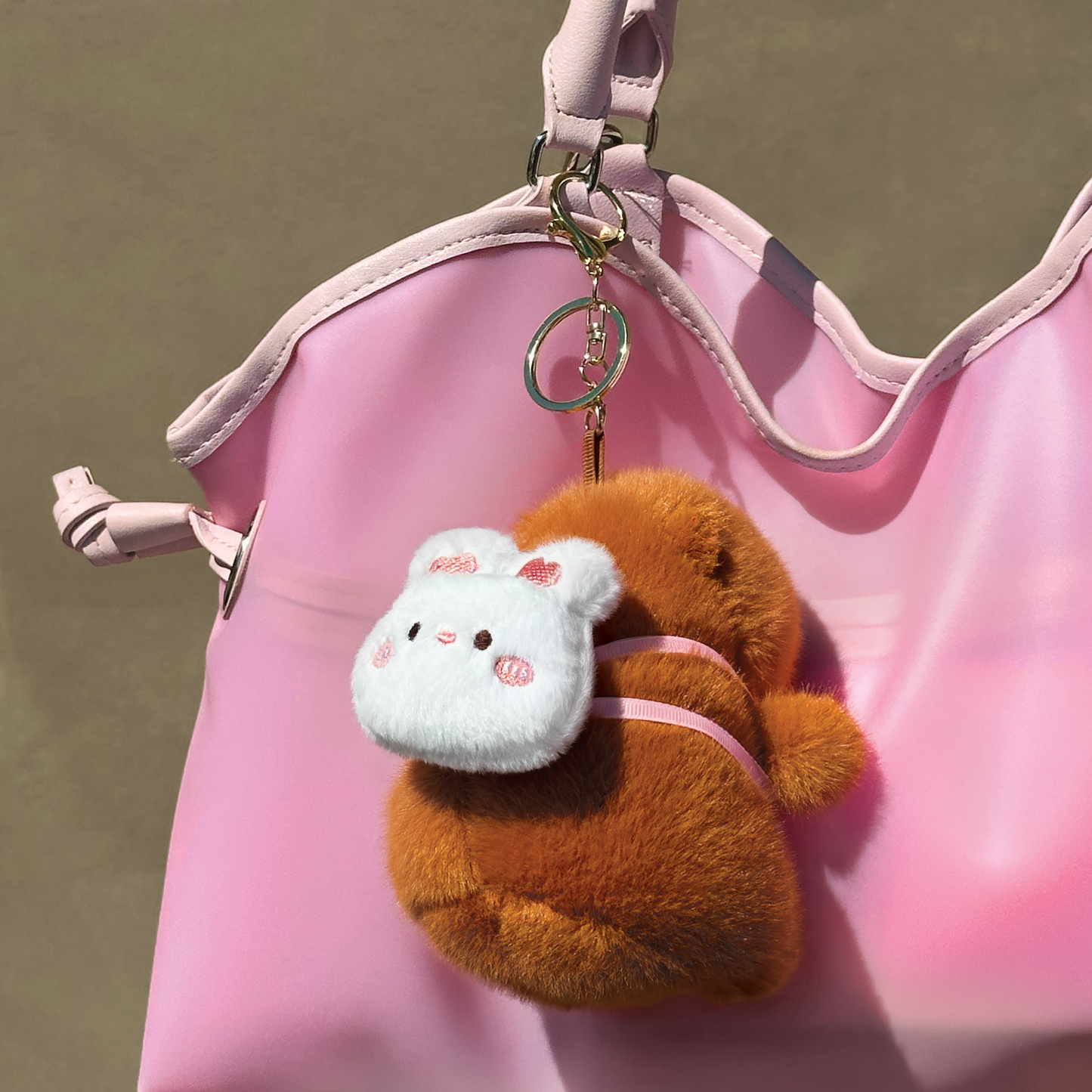 Soft capybara plush keychain wearing a rabbit plush shaped backpack with pink straps and a gold keyring keychain hanging from a pink purse