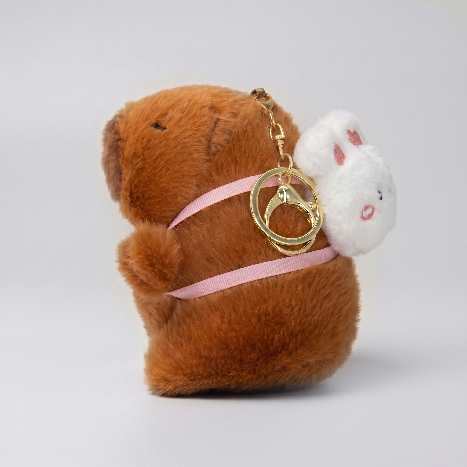 Soft capybara plush keychain wearing a rabbit plush shaped backpack with pink straps and a gold keyring keychain 