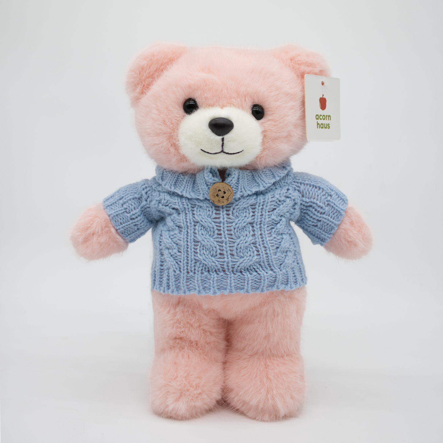 Pink Carrington teddy bear standing upright wearing a blue sweater with one button on a white background