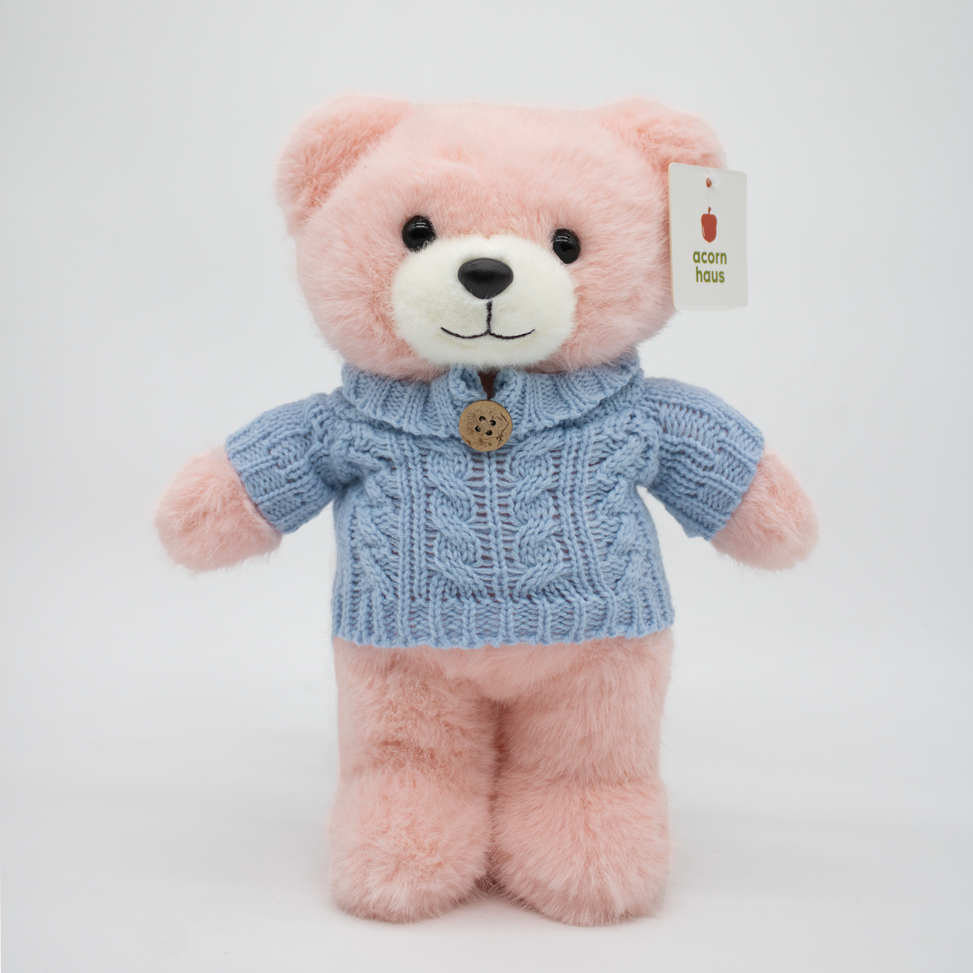 Pink Carrington teddy bear standing upright wearing a blue sweater with one button on a white background