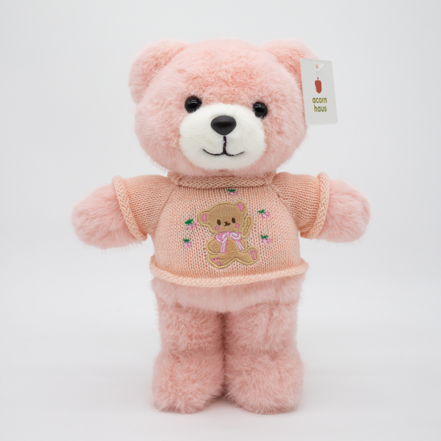 Pink Carrington teddy bear standing upright wearing a beige sweater with a brown bear wearing pink bow design on a white background