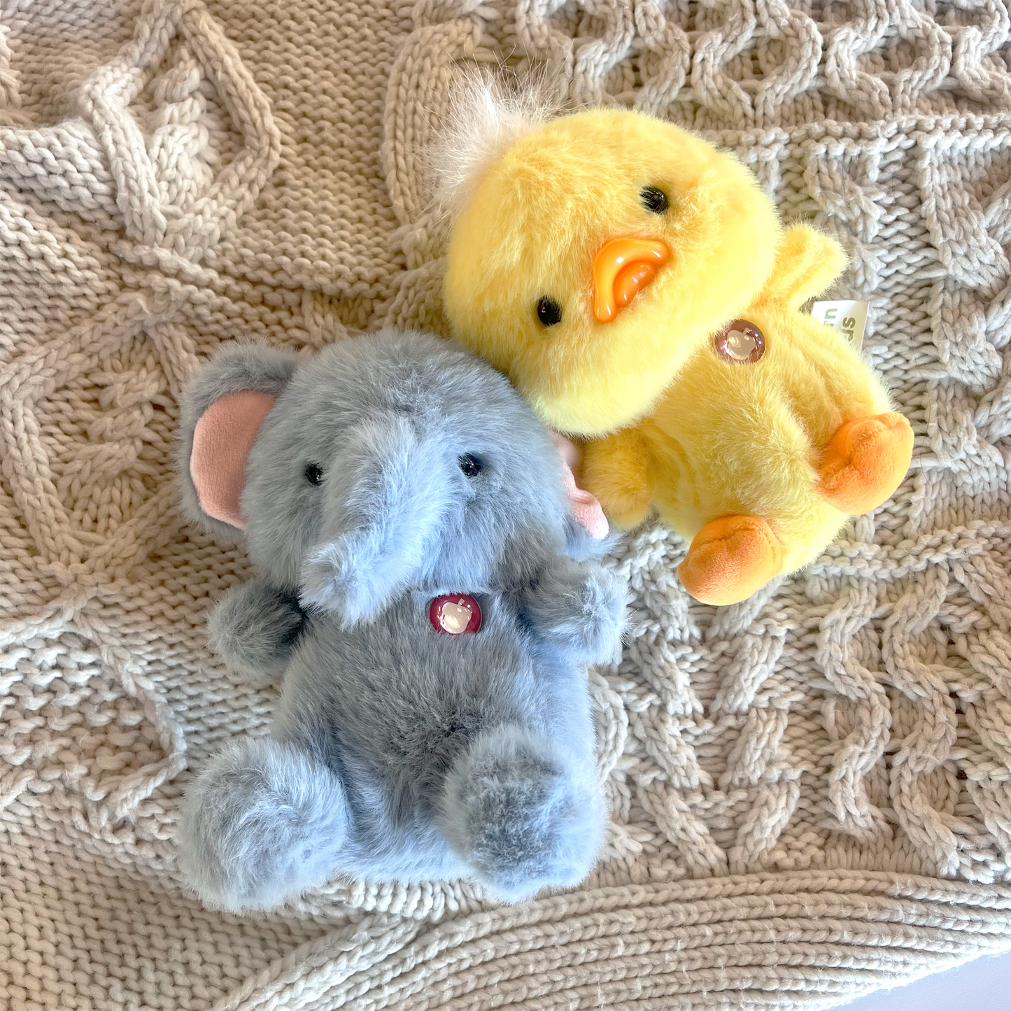 Top-down view of Elephant Puff Little beside Duck Puff Little resting on beige blanket in lifestyle setting