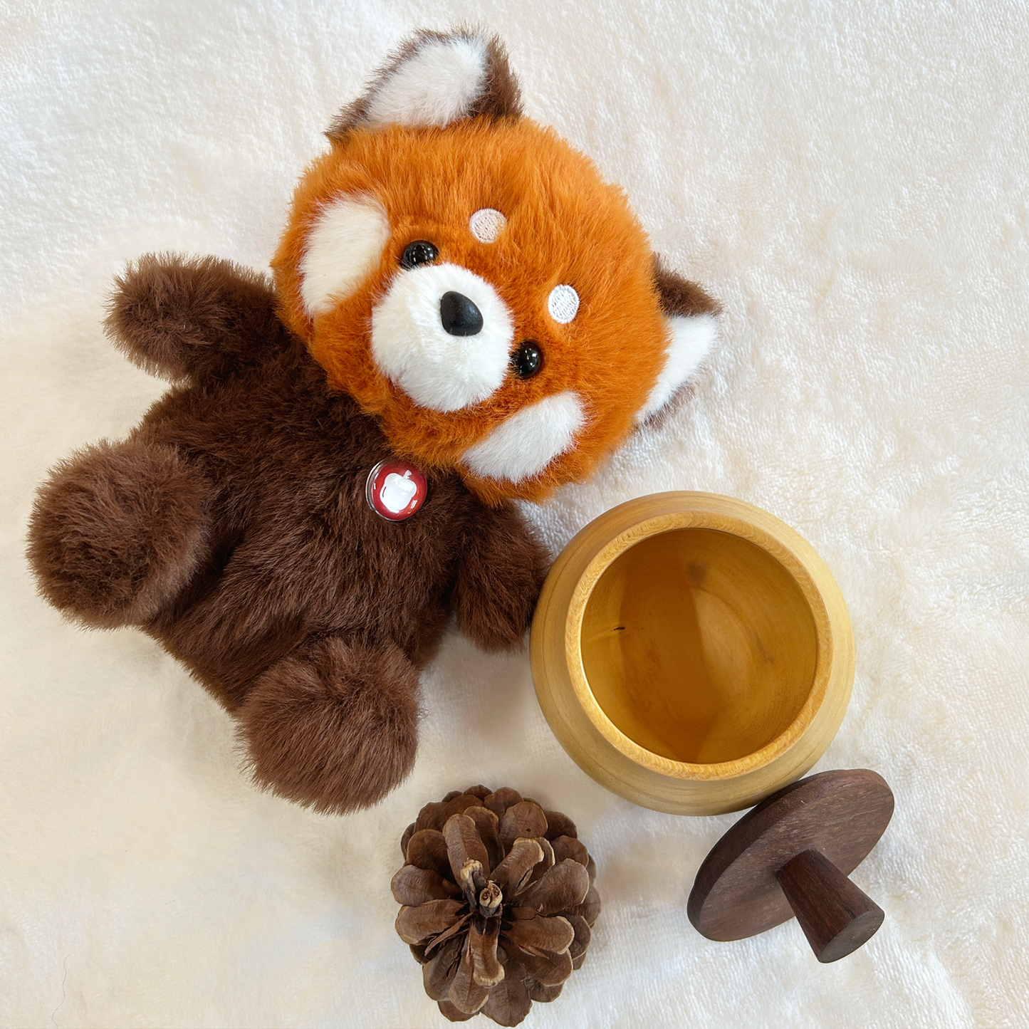 Plush red panda with a wooden acorn and pine cone on a soft white background